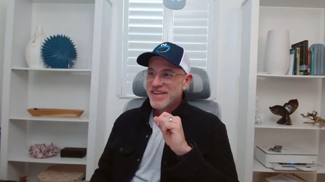 speaker in white shirt facing camera speaking into a microphone with bookshelf behind him